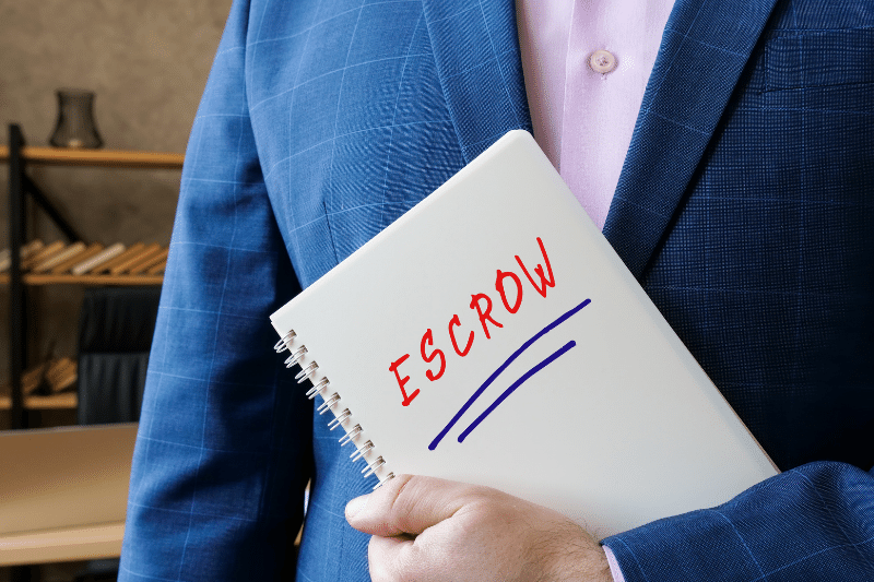 A person in a blue suit holds a notebook with the word ESCROW written in red marker, underlined in blue. The background is an office setting with shelves and hangers.