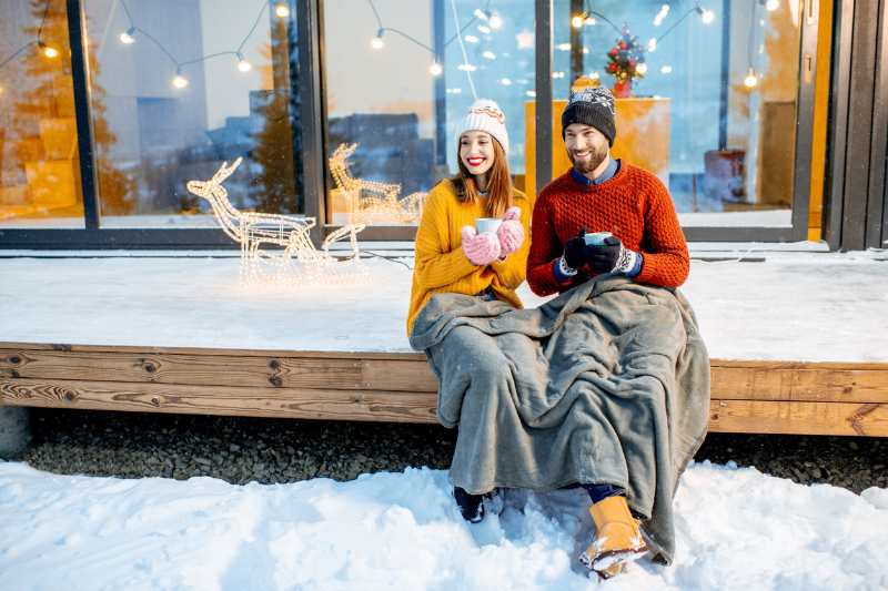 A smiling couple in winter clothes sits on a snowy porch, wrapped in blankets and holding mugs. Festive decorations and string lights are behind them, along with a lit reindeer figure.