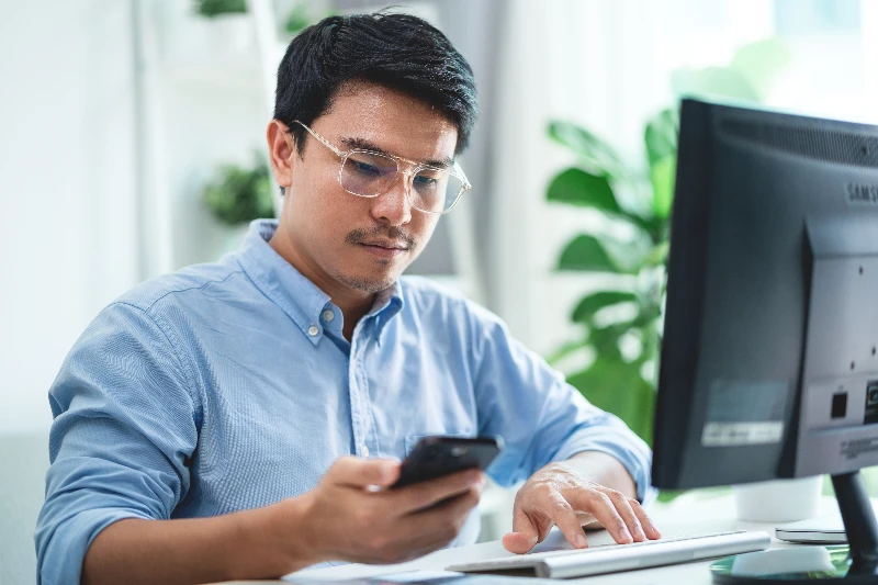 A men wearing glasses is sitting at a desk with a computer and a cell phone